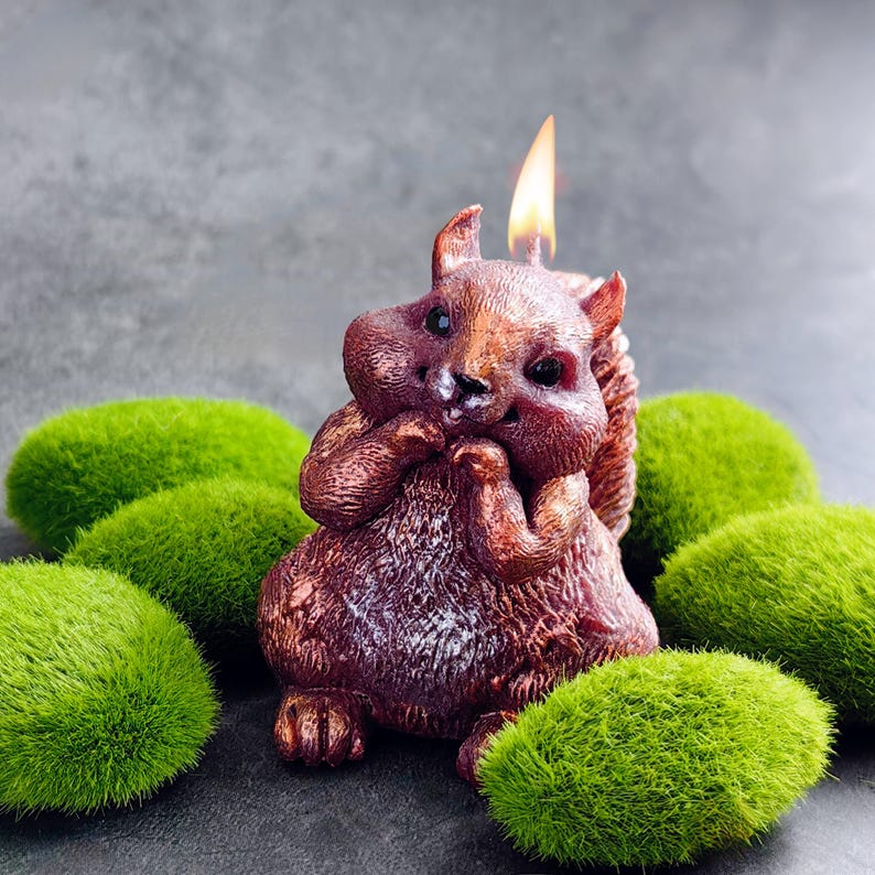 Puede incluir: Una vela de ardilla de color marr&oacute;n y bronce con una mecha encendida. La vela est&aacute; sentada sobre un lecho de musgo verde.