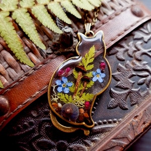 May include: A gold cat-shaped pendant with a clear resin insert containing pressed flowers, ferns, and mushrooms. The pendant is on a brown leather surface.