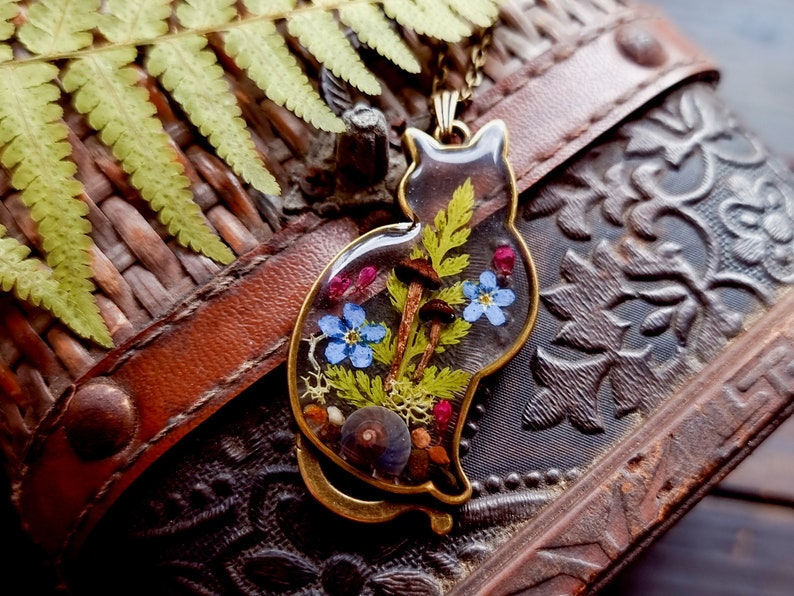 May include: A gold cat-shaped pendant with a clear resin insert containing pressed flowers, ferns, and mushrooms. The pendant is on a brown leather surface.
