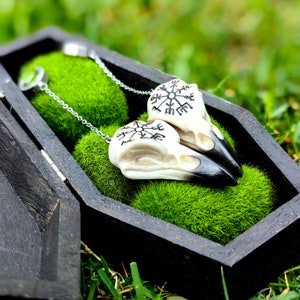 Puede incluir: Un par de colgantes de calavera de cuervo con picos negros y marcas negras intrincadas, exhibidos en una caja negra en forma de ataúd llena de musgo verde. Los colgantes cuelgan de cadenas plateadas.