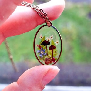 May include: A delicate oval pendant necklace with a clear resin setting featuring pressed flowers, mushrooms, and leaves. The pendant is on a bronze chain.