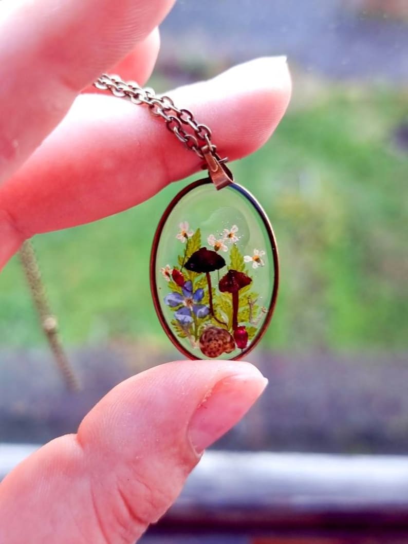 May include: A delicate oval pendant necklace with a clear resin setting featuring pressed flowers, mushrooms, and leaves. The pendant is on a bronze chain.
