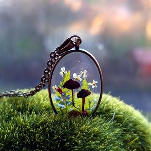 May include: A delicate necklace featuring a clear oval pendant with two small black mushrooms, white flowers, and green leaves encased in resin. The pendant hangs from a silver chain.