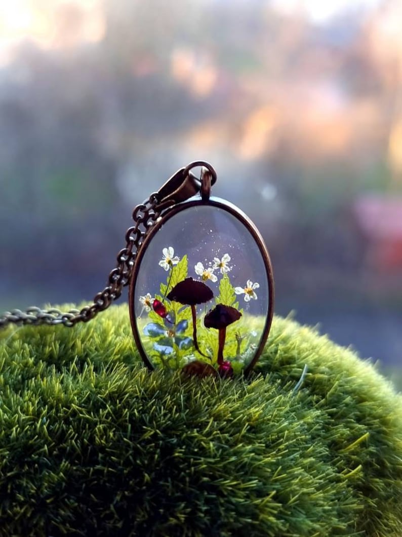 May include: A delicate necklace featuring a clear oval pendant with two small black mushrooms, white flowers, and green leaves encased in resin. The pendant hangs from a silver chain.