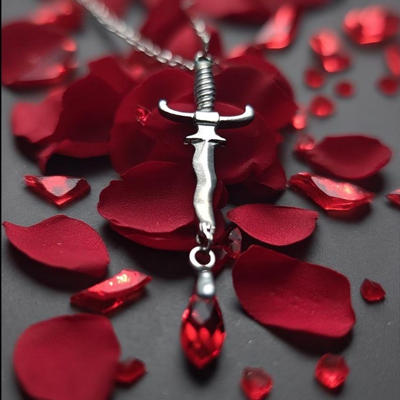 May include: A silver pendant necklace featuring a sword design with a red gemstone at the bottom. The necklace is displayed on a bed of red rose petals, creating a romantic and elegant aesthetic.
