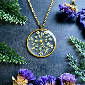 Op de afbeelding: Een gouden ketting met een ronde hanger met een delicate witte bloem geperst in helder hars.