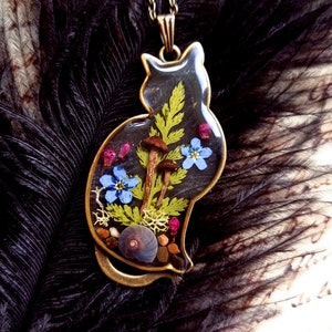 May include: A gold cat-shaped pendant necklace with a clear resin insert containing pressed flowers, ferns, mushrooms, and a snail shell.