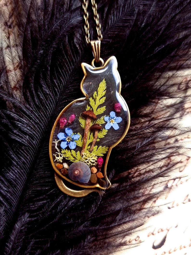 May include: A gold cat-shaped pendant necklace with a clear resin insert containing pressed flowers, ferns, mushrooms, and a snail shell.