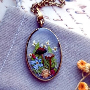 May include: A delicate oval pendant necklace with a clear resin setting featuring pressed flowers and mushrooms. The pendant is made of brass and hangs from a chain.