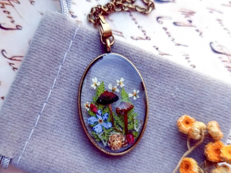 May include: A delicate oval pendant necklace with a clear resin setting featuring pressed flowers and mushrooms. The pendant is made of brass and hangs from a chain.
