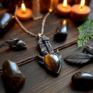 Könnte beinhalten: Eine silberfarbene Halskette mit einem detaillierten Burgdesign und einem großen, ovalen Tigerauge-Stein. Die Halskette wird auf einer dunklen Holzoberfläche präsentiert, umgeben von anderen dekorativen Gegenständen und brennenden Kerzen.