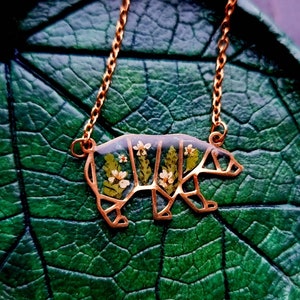 May include: A gold necklace featuring a bear-shaped pendant. The pendant is filled with pressed flowers and leaves, creating a delicate and intricate design.