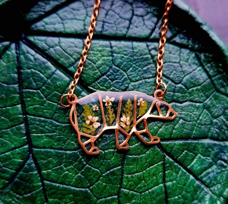 May include: A gold necklace featuring a bear-shaped pendant. The pendant is filled with pressed flowers and leaves, creating a delicate and intricate design.