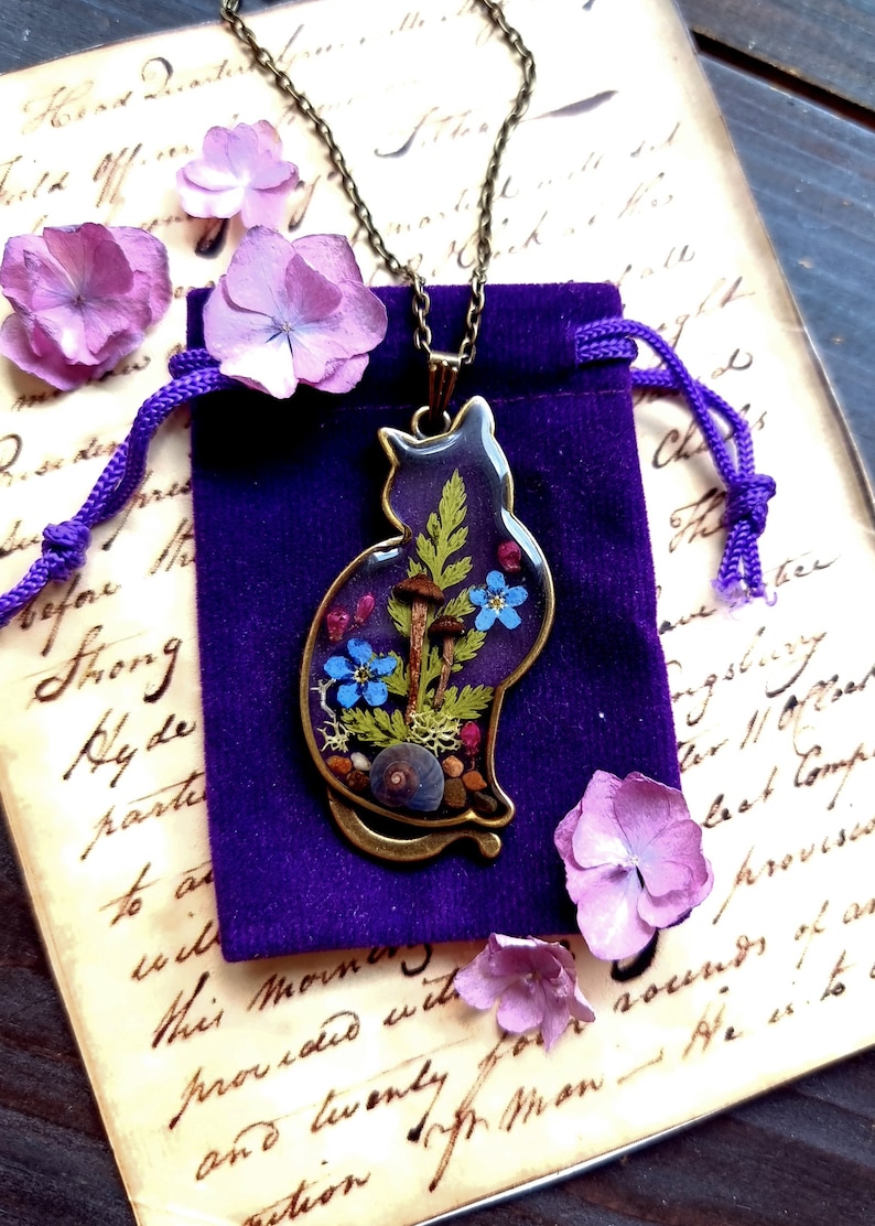 May include: A purple cat-shaped resin pendant necklace with a bronze chain. The pendant contains pressed flowers, leaves, and mushrooms. The necklace is displayed on a purple velvet pouch.