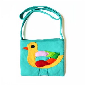 May include: A teal crossbody bag with a yellow duck applique. The duck has a red beak, black eye, and colorful wing details in pink, green, and orange. The bag has a matching teal strap and a zipper closure.
