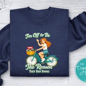 May include: Navy blue sweatshirt with a graphic of a woman riding a teal bicycle with a basket of books. The text reads "I'm Off to Be The Reason They Ban Books" with "1984" on a book in the basket. The Tiger's Trunk LLC logo is on the bottom right.