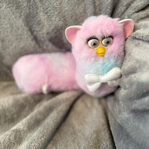 May include: A long, plush toy resembling a Furby with pastel pink, blue, and white tie-dye fur. The creature features large plastic eyes with black pupils, a yellow beak, and two pink ears. A small white fabric bow tie is attached at the neck.
