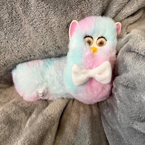 May include: A long, plush toy featuring a pastel tie-dye pattern of pink, blue, and white fur. The creature has large, expressive eyes with dark lashes, small pink-lined ears, and a white fabric bow tie attached to its chest area.