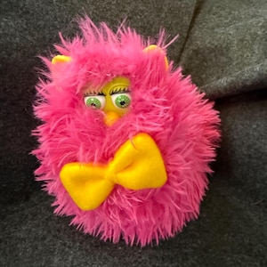 May include: Pink fuzzy toy with green eyes and a yellow bow. The toy has a round shape and is made of soft, plush material.