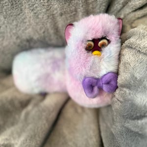 May include: A long, plush toy with tie-dye fur in shades of pink, blue, and white. The toy features large, expressive plastic eyes, a small yellow beak, dark purple inner ears, and a solid purple fabric bow tie attached at the neck area.