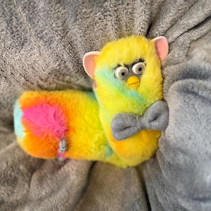 May include: A long, plush toy with vibrant, multi-colored fur in shades of yellow, pink, blue, and orange. The toy features a yellow beak, large white eyes with black pupils, pink inner ears, and a soft, grey fabric bow tie attached at the neck.