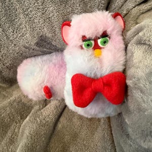 May include: A plush toy featuring a long, soft body with pastel pink, blue, and white fur. The toy has a plastic face with green eyes, a yellow beak, and red ears. A large, bright red fabric bow tie is attached to the front of the chest area.