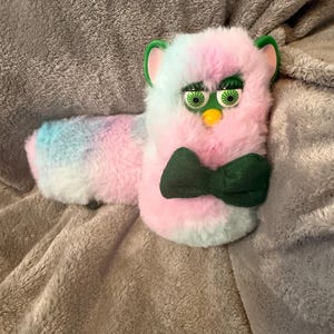 May include: A plush toy featuring soft, pastel pink and blue tie-dye fur, green plastic ears, and green eyes with long lashes. The toy has a small yellow beak and wears a dark green fabric bow tie attached to the front of its fuzzy body.