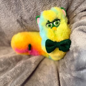May include: A long, plush toy with a Furby-like face featuring green eyes, long eyelashes, and pink inner ears. The body is covered in multi-colored faux fur in shades of yellow, blue, and pink, and it wears a dark green felt bow tie.