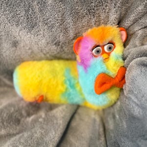 May include: A long, plush toy with tie-dye fur in shades of yellow, blue, pink, and orange. The toy features a Furby-style face with large white eyes, black pupils, orange ears, and a prominent orange bow tie attached to the front of its body.