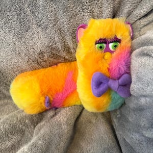 May include: A plush toy featuring a long, multi-colored body with yellow, orange, pink, and light blue faux fur. The head has green eyes with long black eyelashes, purple inner ears, a yellow beak, and a purple felt bow tie at the neck.