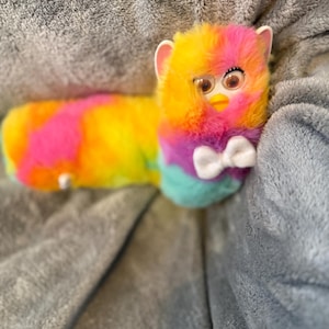 May include: A long, plush toy shaped like a worm with a Furby-style face. The body features a vibrant tie-dye pattern of yellow, orange, pink, purple, and teal. It has large plastic eyes, a yellow beak, white ears, and a white bow tie at the neck.