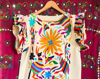 May include: An off-white dress featuring vibrant, multi-colored embroidery of stylized animals and large floral patterns. The garment includes short, ruffled sleeves with matching colorful embroidery and is displayed on a wooden hanger.