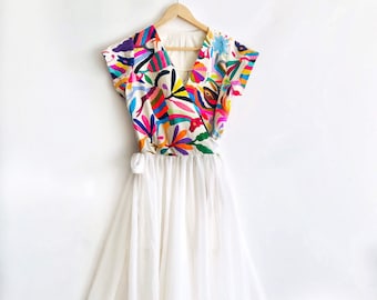May include: A white wrap dress with a colorful embroidered bodice. The bodice features a variety of floral and geometric patterns in shades of pink, blue, green, yellow, and orange. The skirt is a simple white chiffon.
