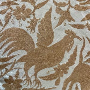 May include: A white fabric featuring intricate tan embroidery of roosters and floral patterns. The design showcases detailed stitching of birds with prominent tail feathers surrounded by various stylized flowers and leaves across the entire surface.