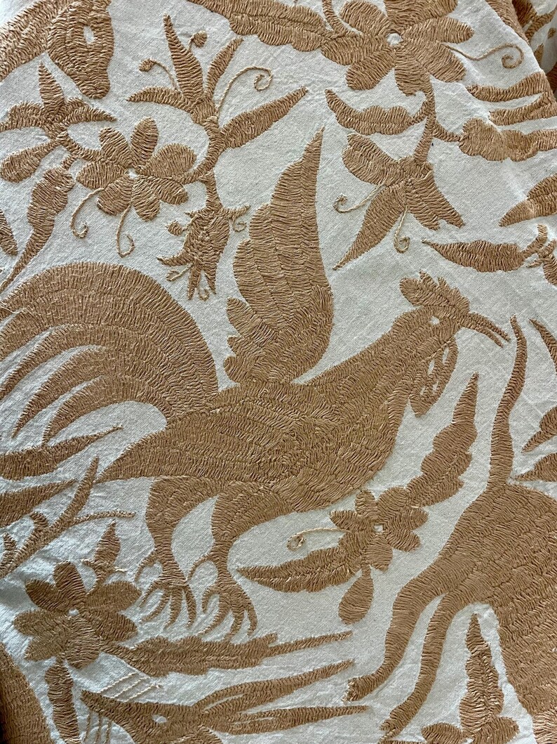 May include: A white fabric featuring intricate tan embroidery of roosters and floral patterns. The design showcases detailed stitching of birds with prominent tail feathers surrounded by various stylized flowers and leaves across the entire surface.