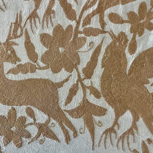 May include: A white fabric featuring intricate tan embroidery depicting a deer, a large bird with spread wings, and various floral elements. The needlework creates a textured, raised pattern across the surface of the textile material.