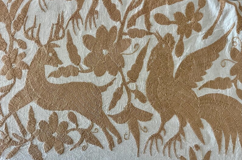 May include: A white fabric featuring intricate tan embroidery depicting a deer, a large bird with spread wings, and various floral elements. The needlework creates a textured, raised pattern across the surface of the textile material.