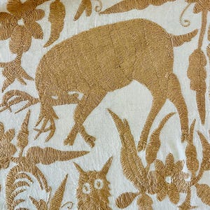 May include: A white textile featuring intricate tan embroidery of a deer grazing among stylized floral and leaf patterns. Below the deer, a small cat-like figure with prominent eyes is depicted, surrounded by dense botanical motifs in tan thread.