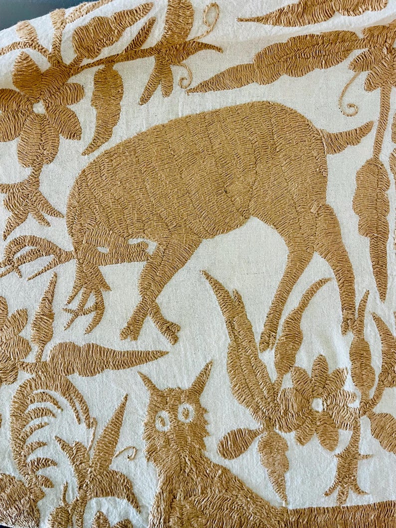May include: A white textile featuring intricate tan embroidery of a deer grazing among stylized floral and leaf patterns. Below the deer, a small cat-like figure with prominent eyes is depicted, surrounded by dense botanical motifs in tan thread.