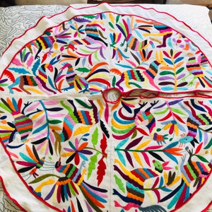 Pode incluir: Uma saia de &aacute;rvore circular em tecido branco com vibrantes padr&otilde;es bordados multicoloridos de animais estilizados e motivos florais. O design inclui uma borda com debrum vermelho e uma abertura central com la&ccedil;os vermelhos.