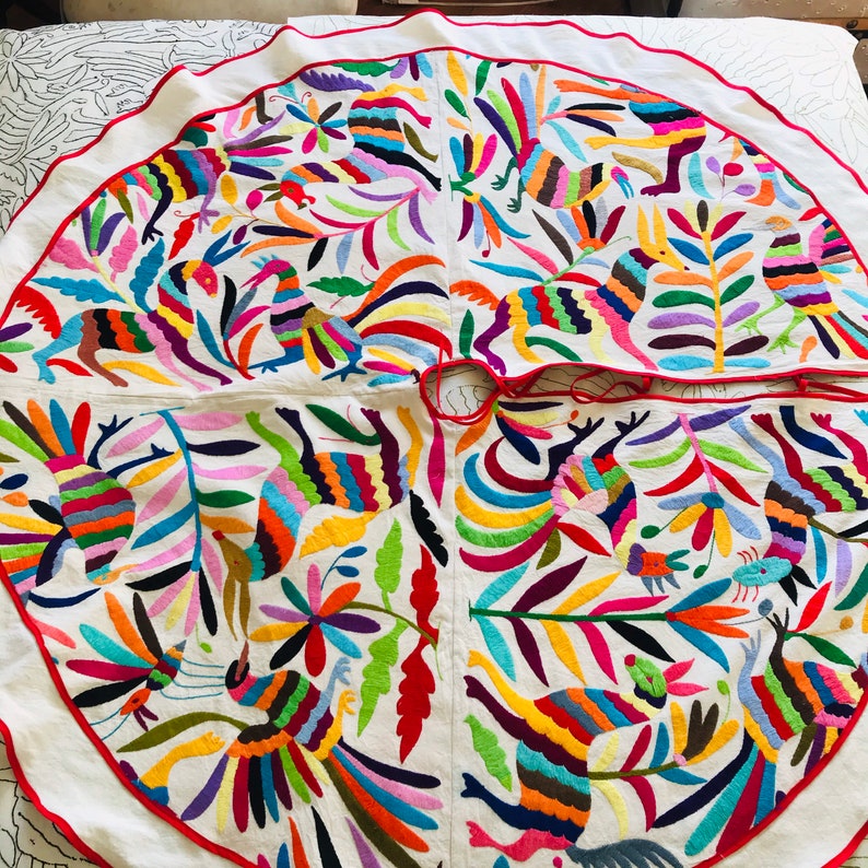 Pode incluir: Uma saia de &aacute;rvore circular em tecido branco com vibrantes padr&otilde;es bordados multicoloridos de animais estilizados e motivos florais. O design inclui uma borda com debrum vermelho e uma abertura central com la&ccedil;os vermelhos.