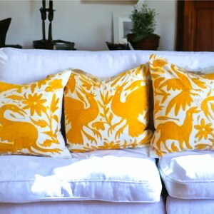 Pode incluir: Três almofadas quadradas com bordados em amarelo vibrante sobre um fundo branco. O design apresenta padrões intrincados de pássaros, flores e folhagens, com cada almofada exibindo uma disposição única destes motivos decorativos.