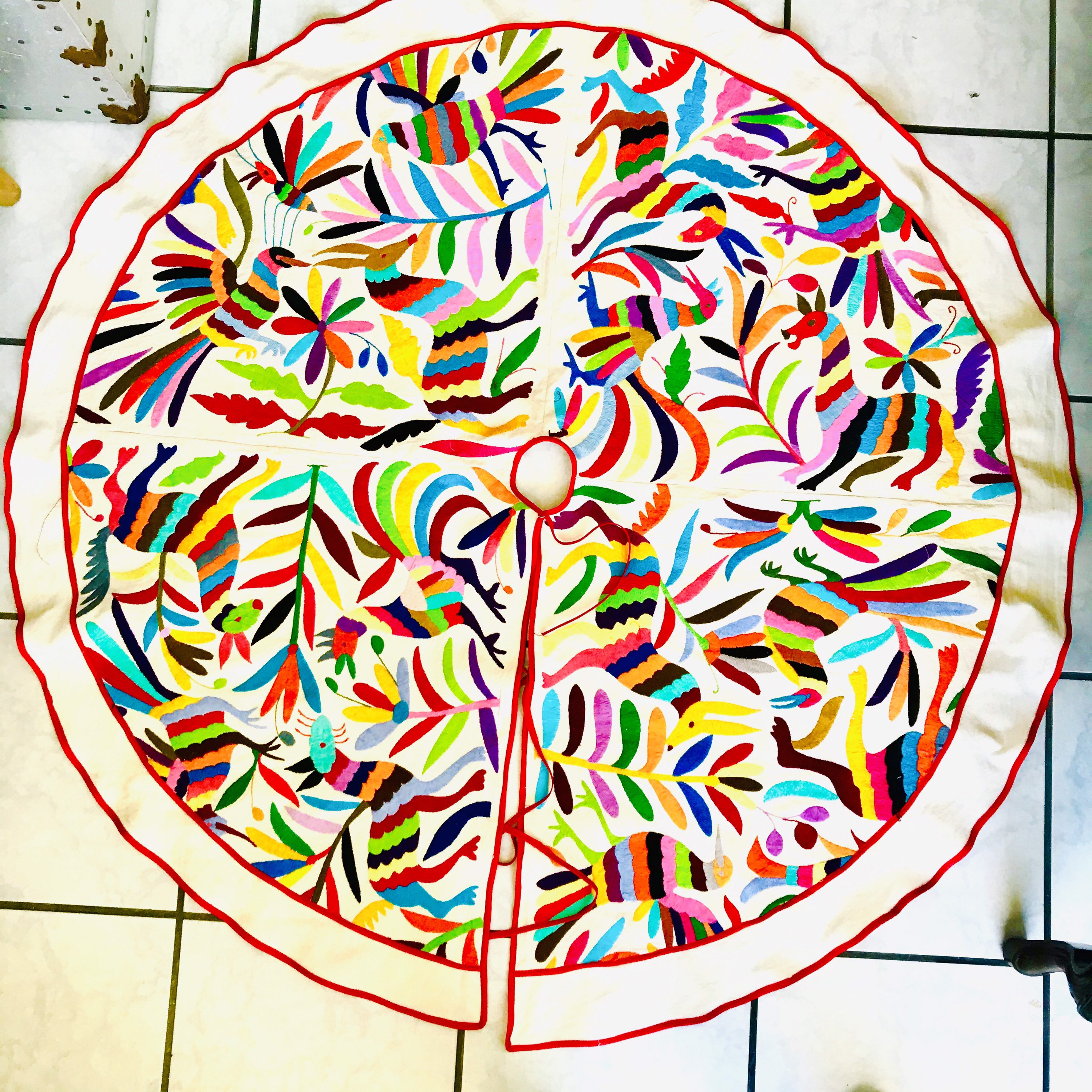 Pode incluir: Uma saia de &aacute;rvore circular em tecido branco sujo com padr&otilde;es bordados multicoloridos vibrantes de p&aacute;ssaros, veados e motivos florais. O design inclui uma abertura central e uma fenda, com uma borda festonada vermelha viva.