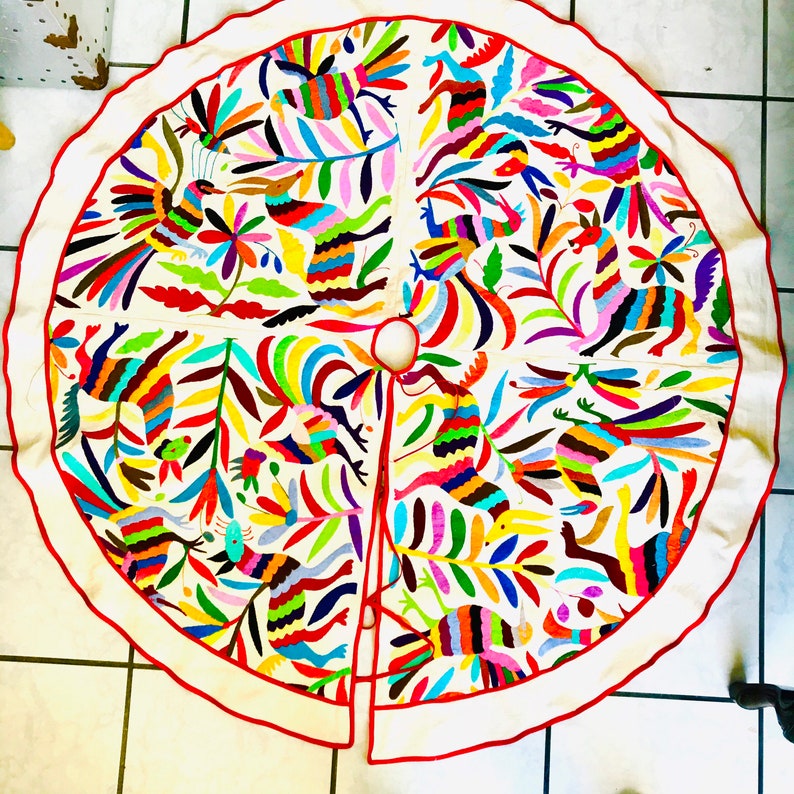 Pode incluir: Uma saia de &aacute;rvore circular em tecido branco sujo com padr&otilde;es bordados multicoloridos vibrantes de p&aacute;ssaros, veados e motivos florais. O design inclui uma abertura central e uma fenda, com uma borda festonada vermelha viva.