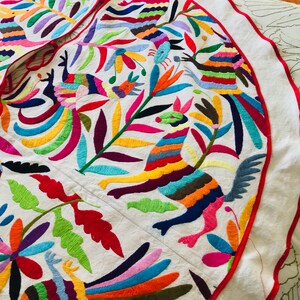 Pode incluir: Uma saia de &aacute;rvore circular de tecido branco com bordados vibrantes e multicoloridos de animais estilizados e padr&otilde;es florais. O design inclui p&aacute;ssaros e criaturas semelhantes a veados em tons de vermelho, azul, amarelo e verde, com uma borda vermelha.
