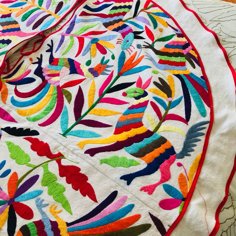 Pode incluir: Uma saia de &aacute;rvore circular de tecido branco com bordados vibrantes e multicoloridos de animais estilizados e padr&otilde;es florais. O design inclui p&aacute;ssaros e criaturas semelhantes a veados em tons de vermelho, azul, amarelo e verde, com uma borda vermelha.