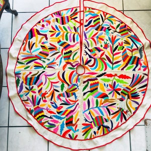 Pode incluir: Uma saia de &aacute;rvore de Natal circular em tom branco sujo com padr&otilde;es vibrantes bordados &agrave; m&atilde;o de p&aacute;ssaros e folhagens em v&aacute;rias cores. O tecido tem uma borda com debrum vermelho e uma abertura central. Texto: Christmas Tree skirt e HAND EMBROIDERY BY ARTE OTOMI.