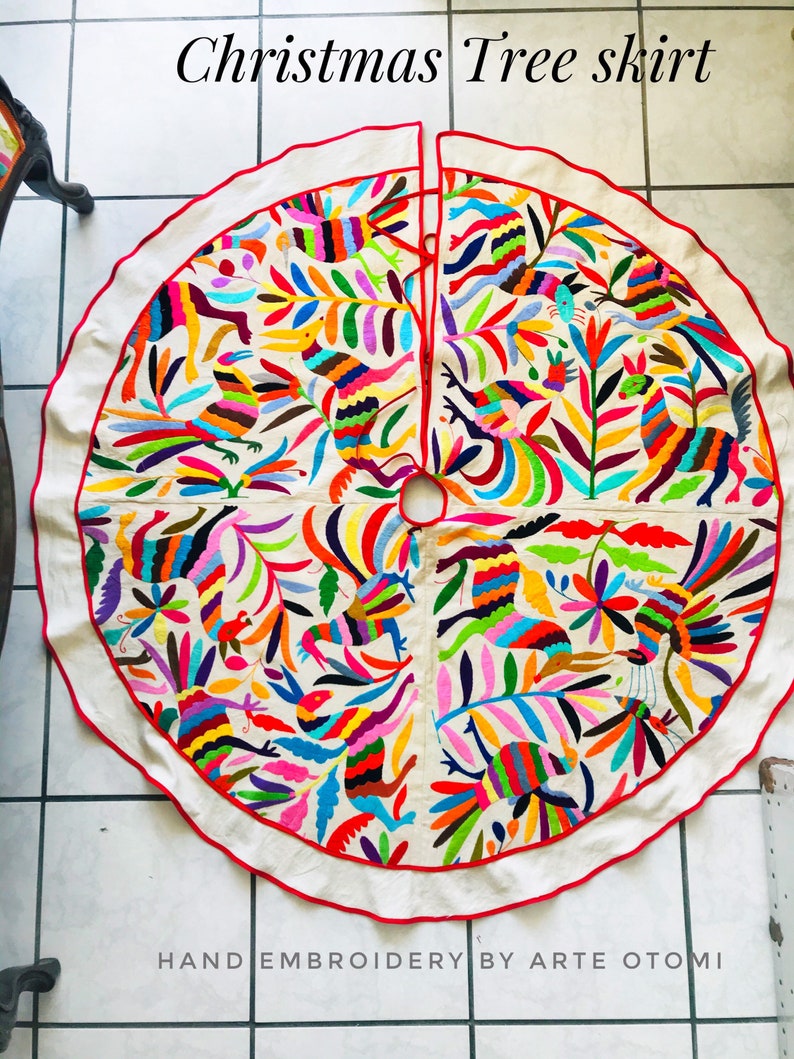 Pode incluir: Uma saia de &aacute;rvore de Natal circular em tom branco sujo com padr&otilde;es vibrantes bordados &agrave; m&atilde;o de p&aacute;ssaros e folhagens em v&aacute;rias cores. O tecido tem uma borda com debrum vermelho e uma abertura central. Texto: Christmas Tree skirt e HAND EMBROIDERY BY ARTE OTOMI.