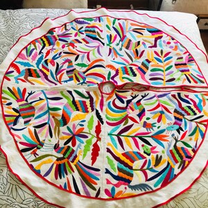Pode incluir: Uma saia de &aacute;rvore circular em tecido branco com padr&otilde;es vibrantes bordados &agrave; m&atilde;o de animais estilizados e motivos florais. O design inclui um rebordo vermelho na extremidade exterior e uma abertura central com la&ccedil;os vermelhos.