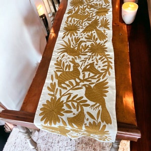 May include: Cream-colored table runner with a gold embroidered design of birds, flowers, and foliage. The runner is displayed on a dark wooden table, with lit candles in the background. The intricate embroidery adds a decorative touch.